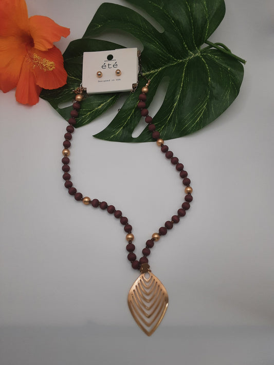 The SPECIAL EFFECTS ETE EMN1400 LEAF necklace, showcasing maroon and gold beads with a gold leaf-shaped pendant, is displayed on a white background adorned with a green leaf and an orange flower.