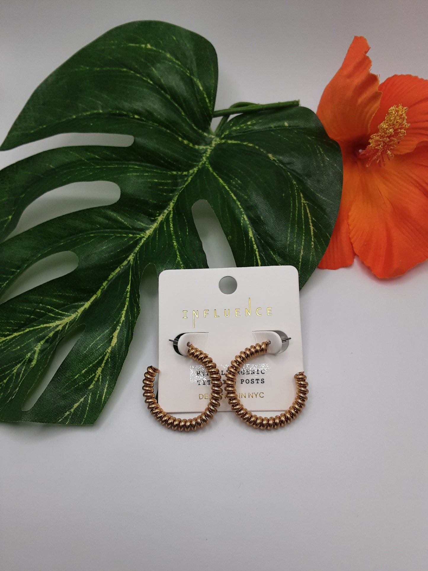 The Curly Metal Wire Hoop Earrings Gold Tone by SPECIAL EFFECTS are displayed on a white card labeled "INFLUENCE," placed on a large green leaf next to a vibrant orange flower. These stunning 1 1/2 inch hoop earrings add a touch of elegance to any outfit.