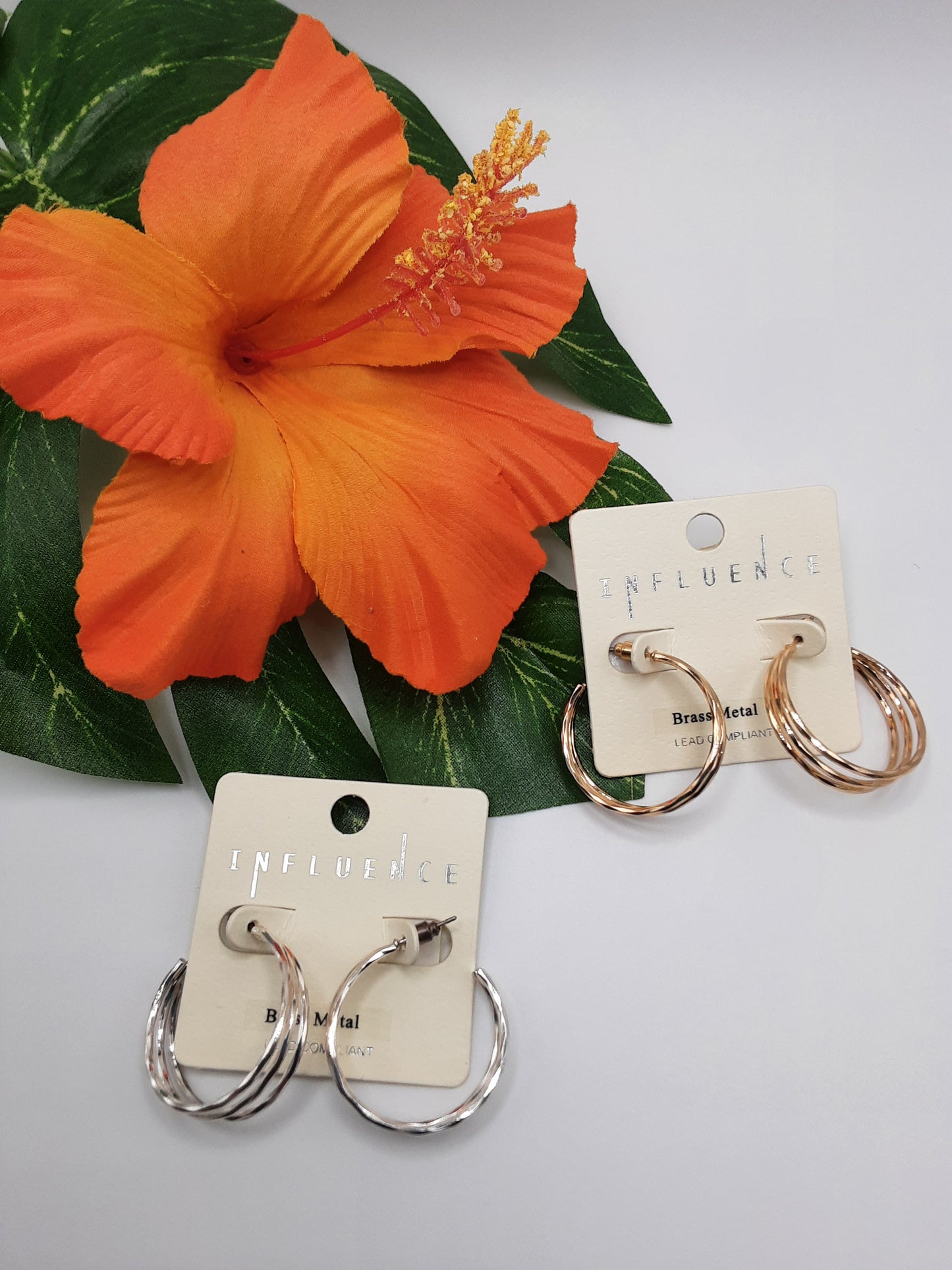 Close-up of two pairs of SPECIAL EFFECTS 3 ROW 30MM HOOP EARRING displayed on label cards, with a large orange hibiscus flower and green leaves in the background.