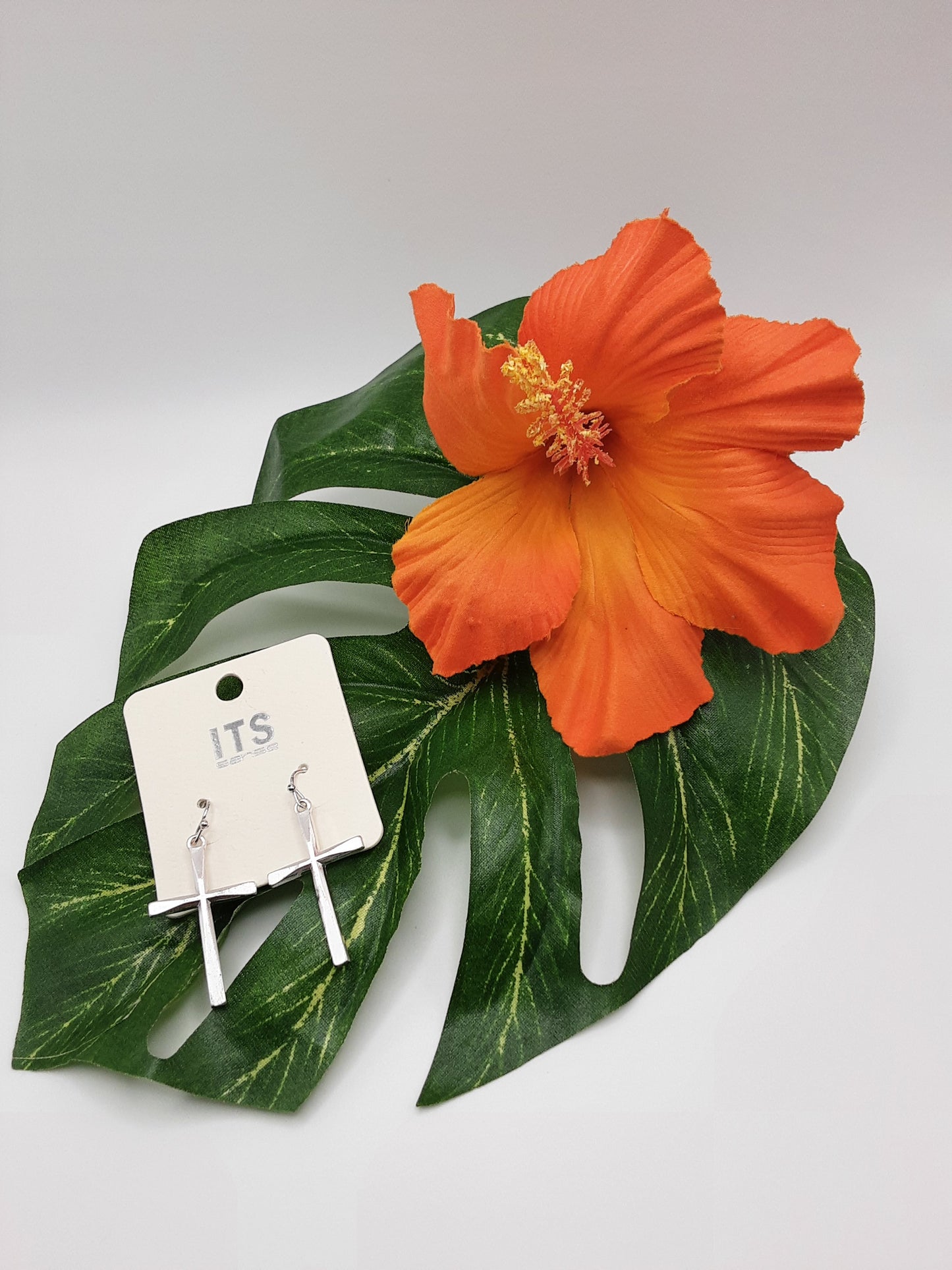 A pair of Fashion Go ITS ME20193 CROSS silver earrings displayed on a white ITS card, set against a green leaf with an orange hibiscus flower.