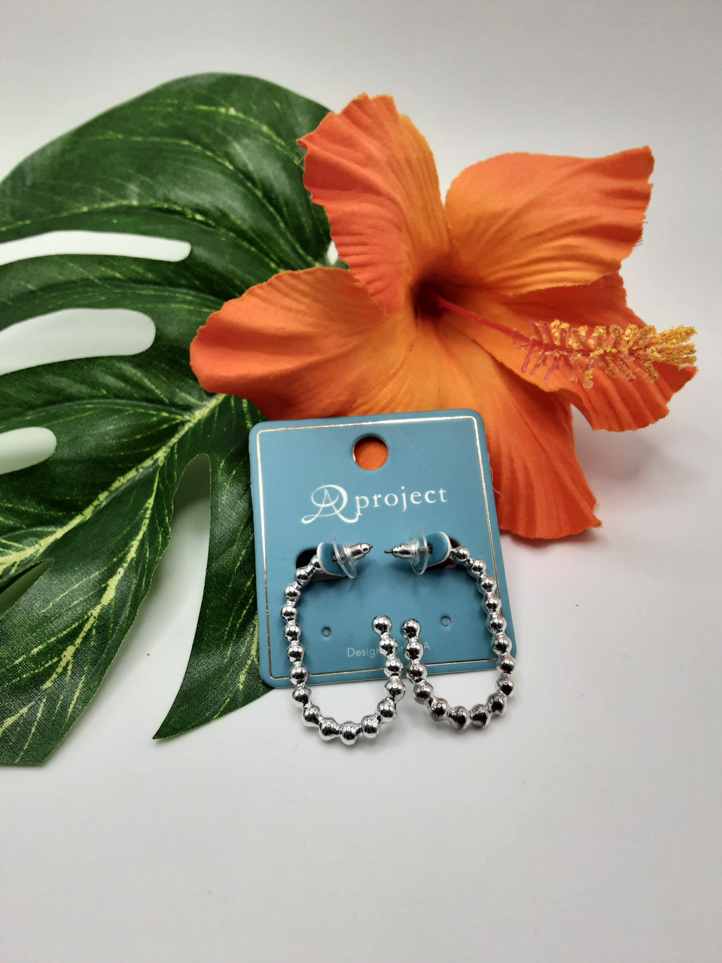 A pair of APROJECT QE2592 BEAD earrings from SPECIAL EFFECTS, displayed on a blue card labeled with the same, are placed in front of a large green leaf and an orange hibiscus flower. Main image