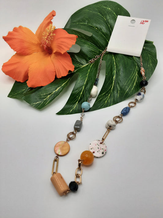 The ANARCHY STREET YYN005ULTI by Fashion Go, a beaded necklace featuring a mix of round and irregularly shaped beads, is artfully displayed on a green leaf alongside an orange hibiscus flower. A visible price tag indicates $12.00.