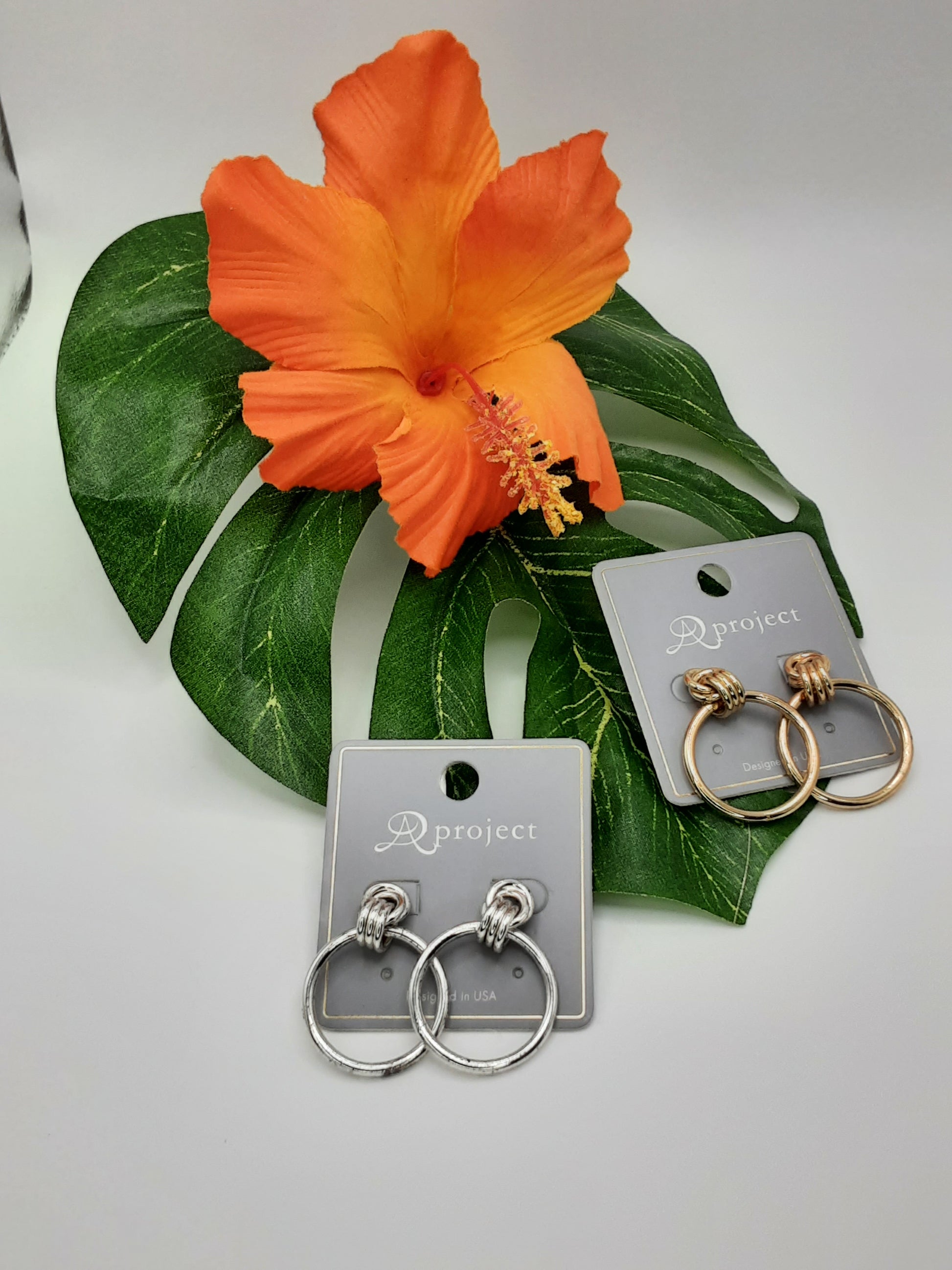 Two pairs of APROJECT QE3390 DOORKNOCKER hoop earrings by SPECIAL EFFECTS are displayed on cards and placed on a green leaf and an orange hibiscus flower. One pair is silver, the other is gold.