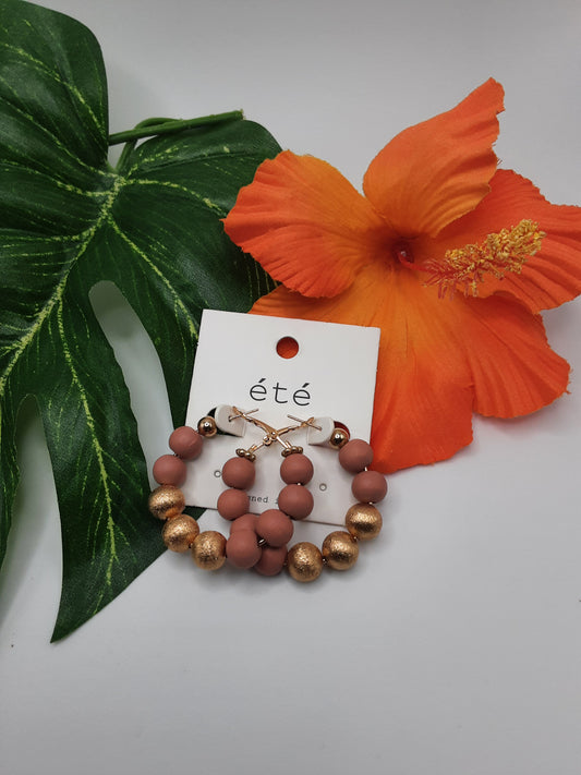 Close-up of SPECIAL EFFECTS ETE TE1230 ASSORTED gold and coral bead hoop earrings displayed on a branded card, set against a backdrop of a large green leaf and an orange hibiscus flower.