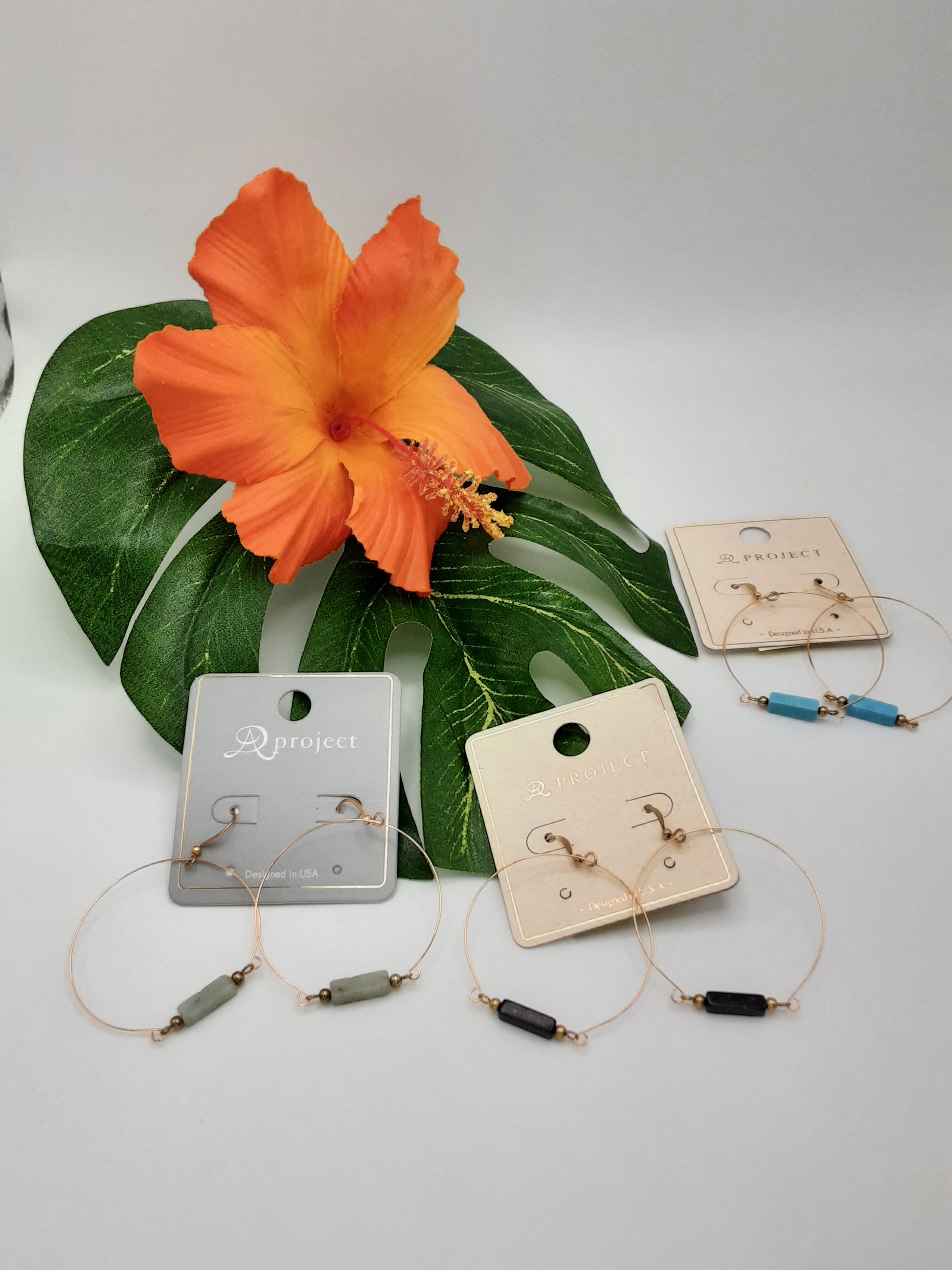 Three pairs of the APROJECT CE3049 ASSORTED earrings by SPECIAL EFFECTS are displayed on cards next to a large green leaf and an orange hibiscus flower. The earrings feature hoop designs with small cylindrical beads in green, black, and blue.