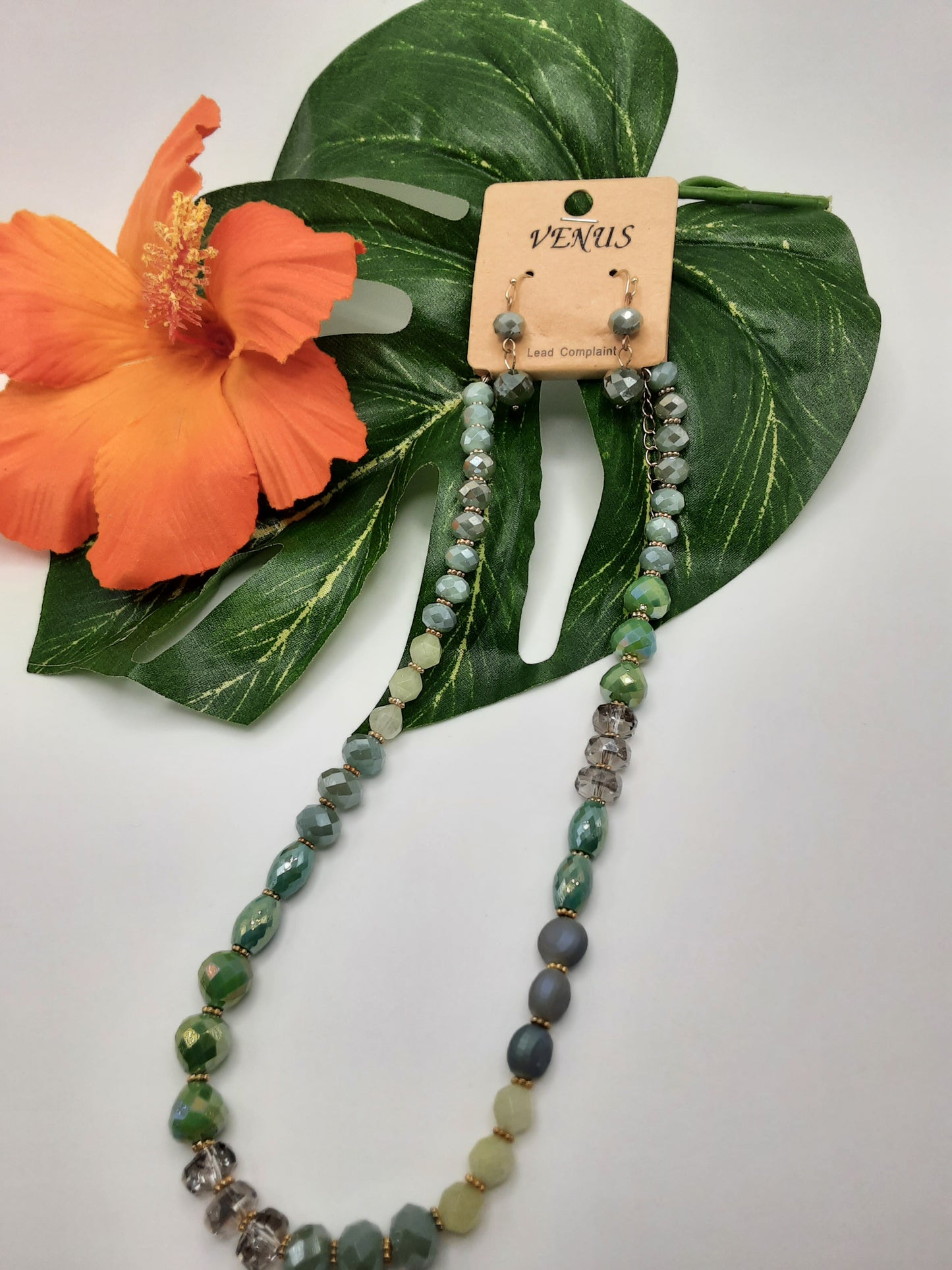 The VENUS NE6642MT CIRCLES beaded necklace and matching earrings from SPECIAL EFFECTS, elegantly displayed on a green leaf alongside an orange hibiscus flower.