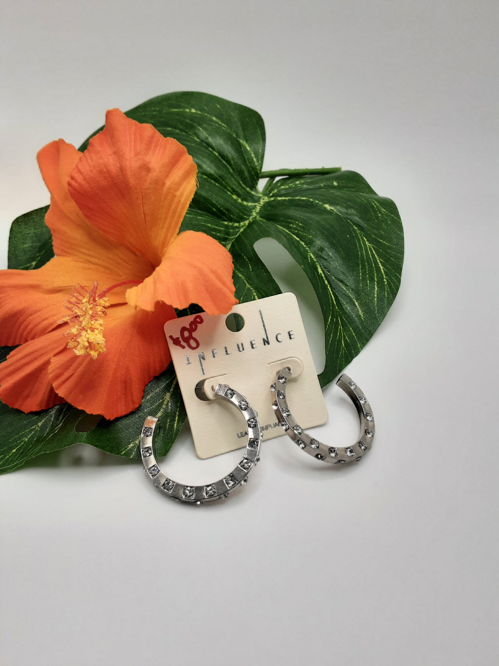 A pair of silver hoop earrings with studded details from SPECIAL EFFECTS displays elegantly on a white tag labeled "INFLUENCE B6E7227 ASSORTED," next to an orange hibiscus flower and a green leaf.