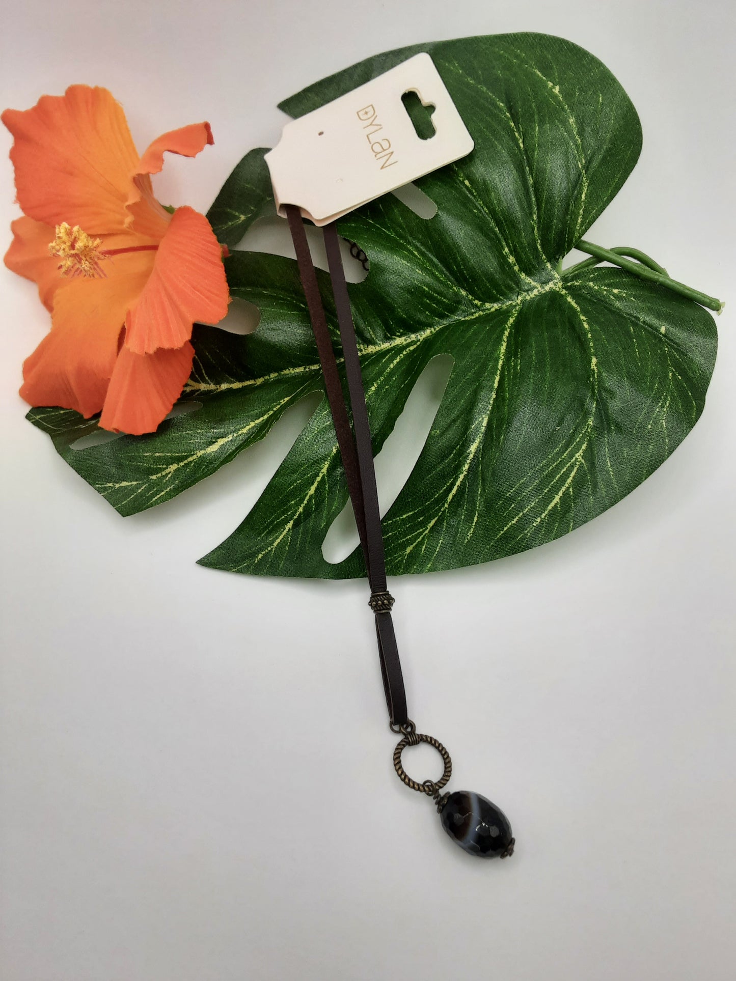 The DYLAN 59093 necklace by Fashion Go, featuring a black pendant, is displayed on a green leaf alongside an orange flower.