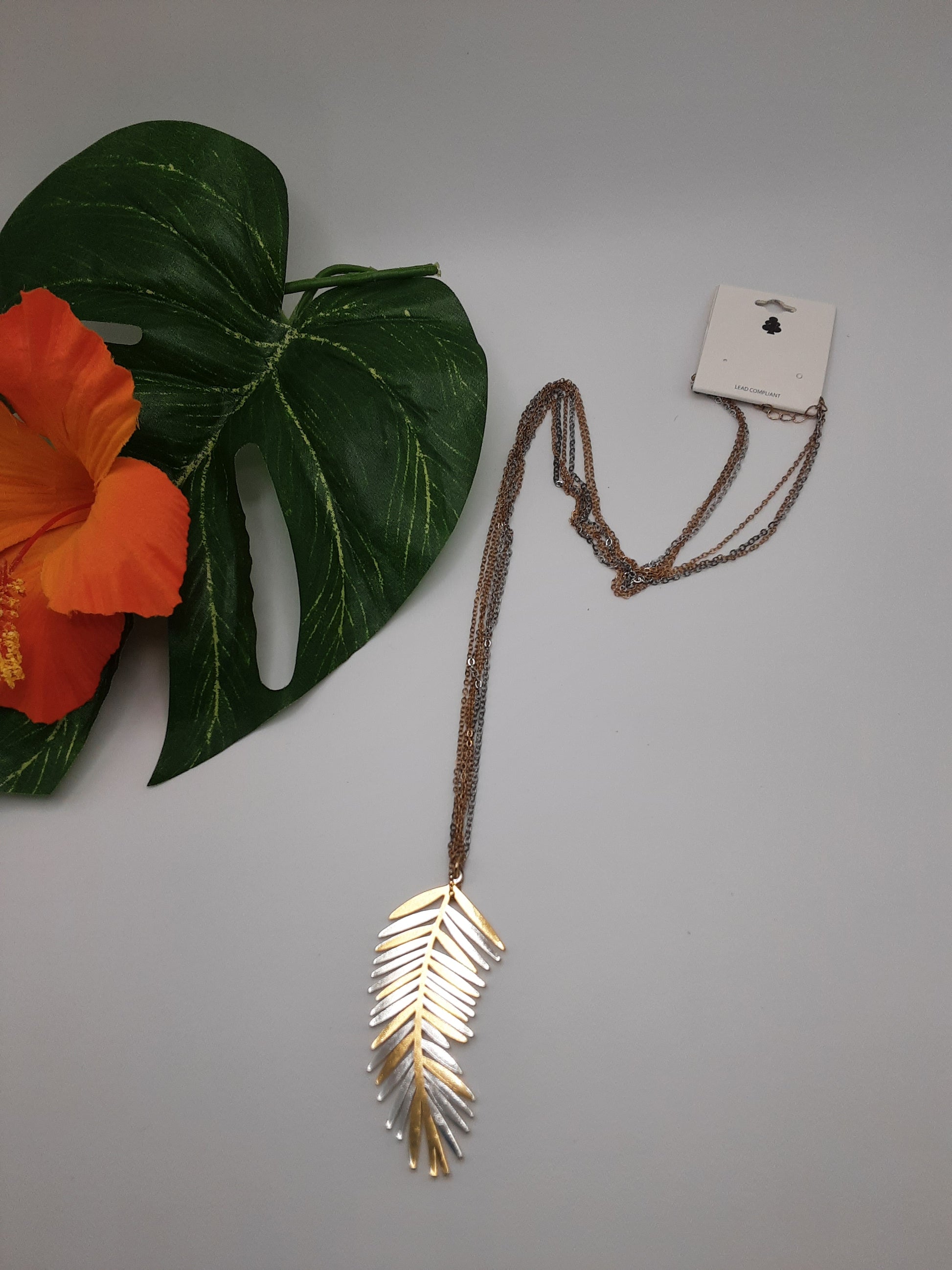 The URBANISTA NW3153 LEAF pendant necklace by Fashion Go, featuring a gold-colored leaf design and long chain, is displayed on a gray background next to a green leaf and an orange flower.