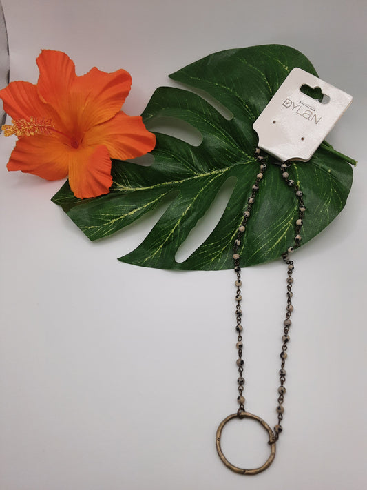 A metal necklace with a circular pendant is displayed on a large green leaf alongside an orange hibiscus flower. The necklace's tag reads "DYLAN 60364 CIRCLE" by Fashion Go.