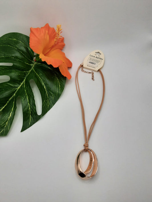 A LOVE ASHLEY 145NK85873 ASSORTED necklace from Fashion Go, featuring a double hoop pendant and brown cord, showcased beside a green leaf and an orange hibiscus flower.