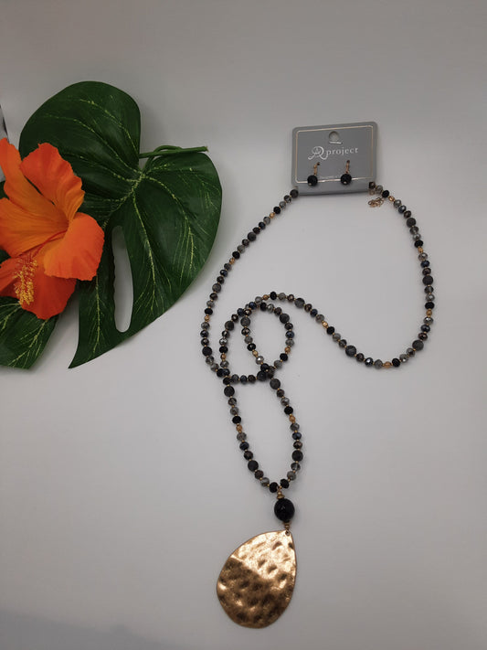 The APROJECT CNE3120 BEADED necklace by SPECIAL EFFECTS, featuring a hammered gold pendant and matching earrings, displayed on a white surface next to a green leaf and an orange flower.