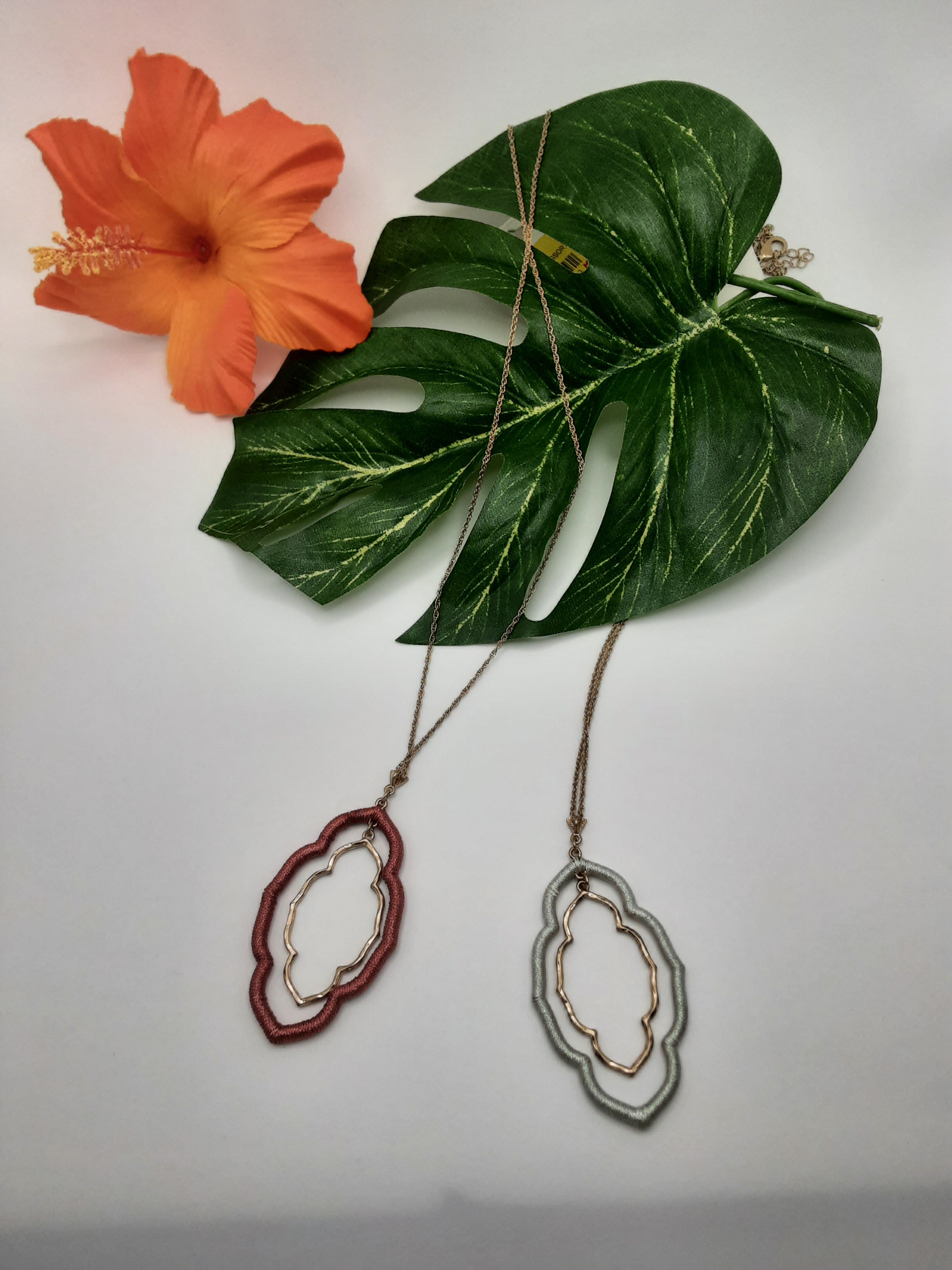 Two URBANISTA NW7150 ASSORTED necklaces by Fashion Go, featuring one with a red geometric pendant and the other with a silver geometric pendant, are displayed on a white surface along with a green leaf and an orange hibiscus flower. Main image
