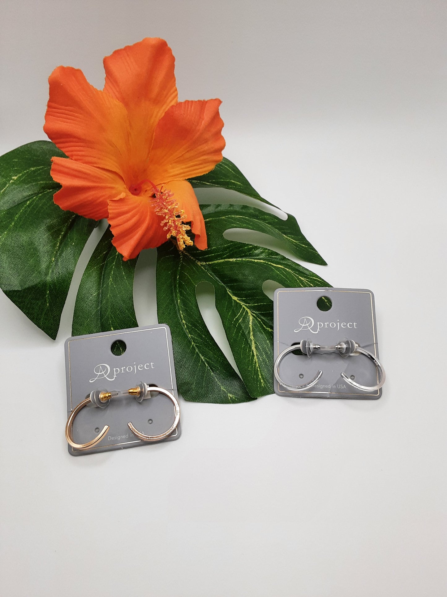 Two pairs of hoop earrings from the APROJECT QE3670 ASSORTED line by SPECIAL EFFECTS, one in gold and one in silver, are showcased on display cards, set against a large green leaf and accented with an orange hibiscus flower.