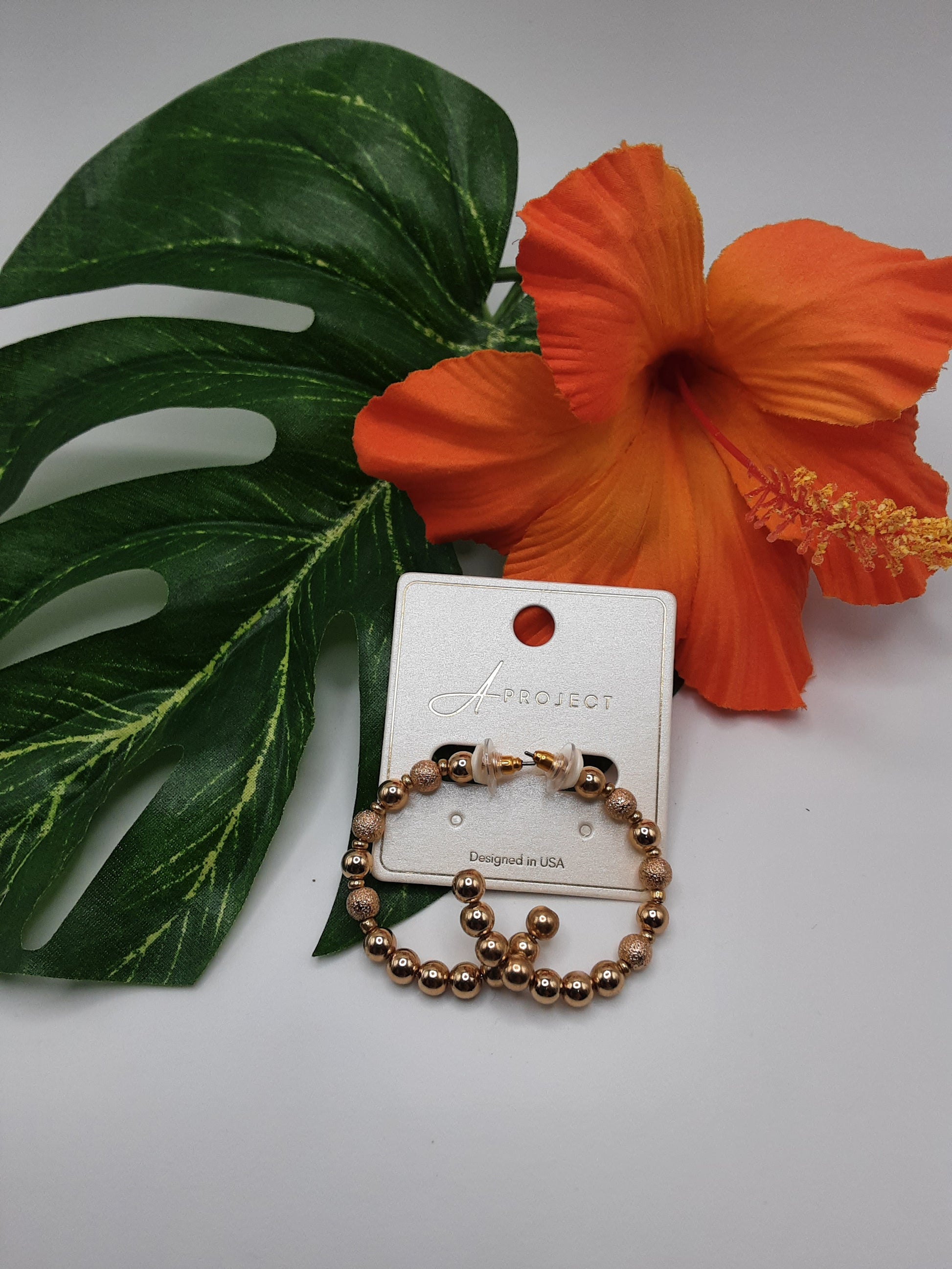 A gold bead bracelet from the "SPECIAL EFFECTS APROJECT KE2297 HOOP" collection is displayed against a white background, adorned with a green leaf and an orange flower.