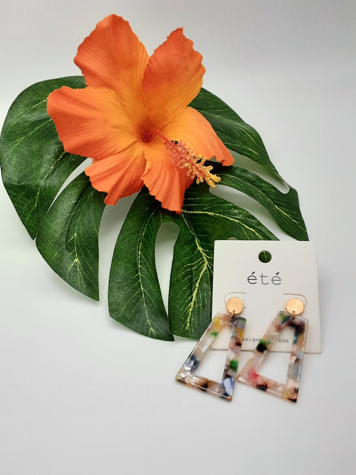 A pair of vibrant Acetate Trapezoid Shaped Earrings from SPECIAL EFFECTS, displayed on a white card labeled "été," placed beside a large, vibrant orange flower and green leaves.