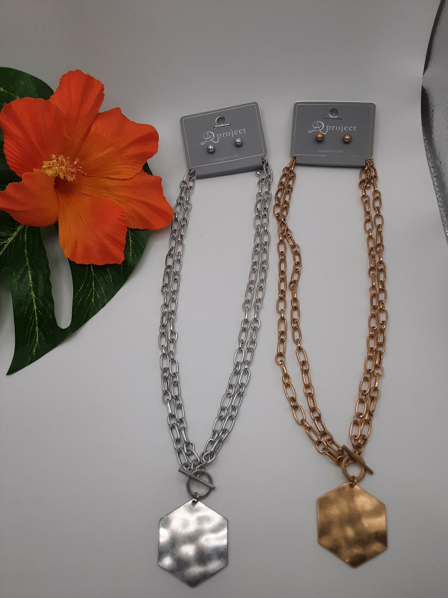 Two SPECIAL EFFECTS chain necklaces, featuring hexagonal pendants from the A PROJECT QNE 2525 HEXAGON collection—one in silver and the other in gold—displayed next to an artificial orange flower on a white surface.