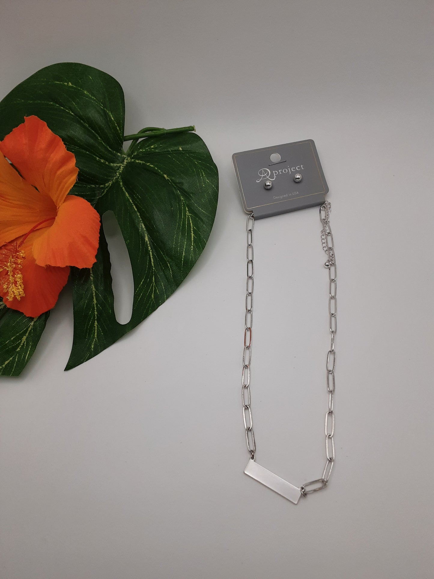 The A PROJECT QNE2741 ASSORTED from SPECIAL EFFECTS, featuring a silver chain necklace with a rectangular pendant, is displayed on a white background with an orange hibiscus flower and green leaves to the side.