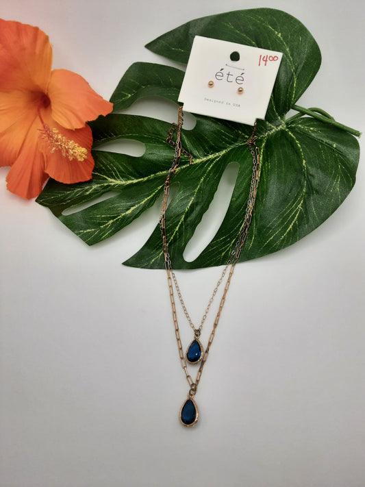 The SPECIAL EFFECTS ETE TNE1036 TEARDROP, a gold-tone layered necklace adorned with blue teardrop pendants and priced at $14.00, is presented on a white tag labeled "ete," showcased beautifully against a green leaf with an orange hibiscus flower.