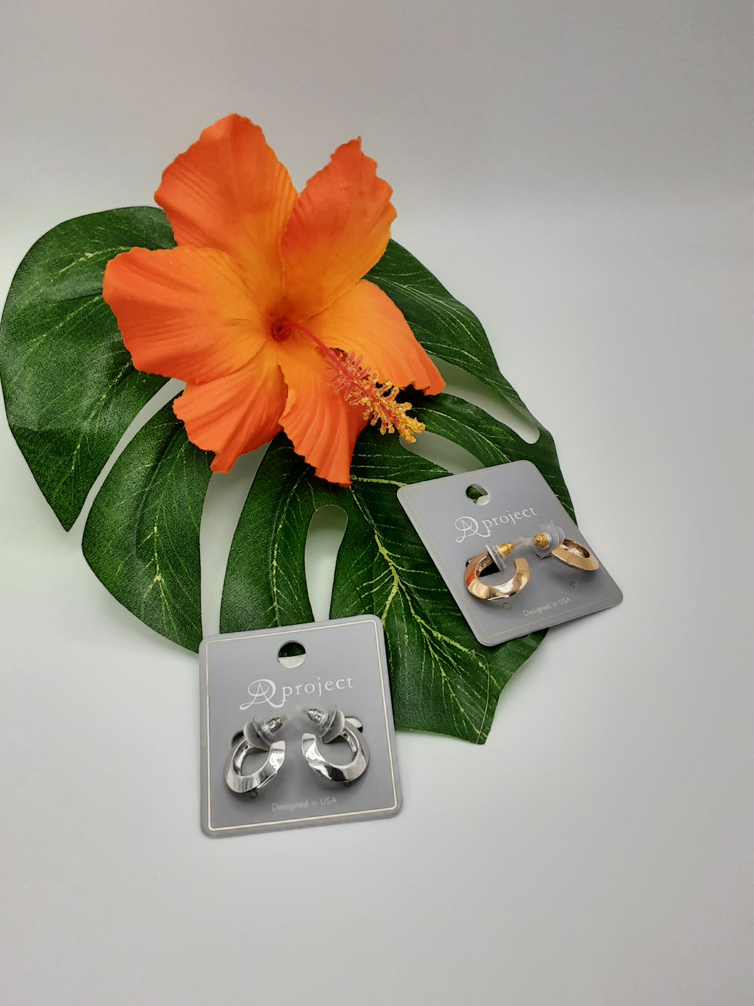 APROJECT 3796 HOOP earrings by SPECIAL EFFECTS, available in gold and silver, are showcased on cards featuring a large orange hibiscus flower and green leaf in the background. Main image