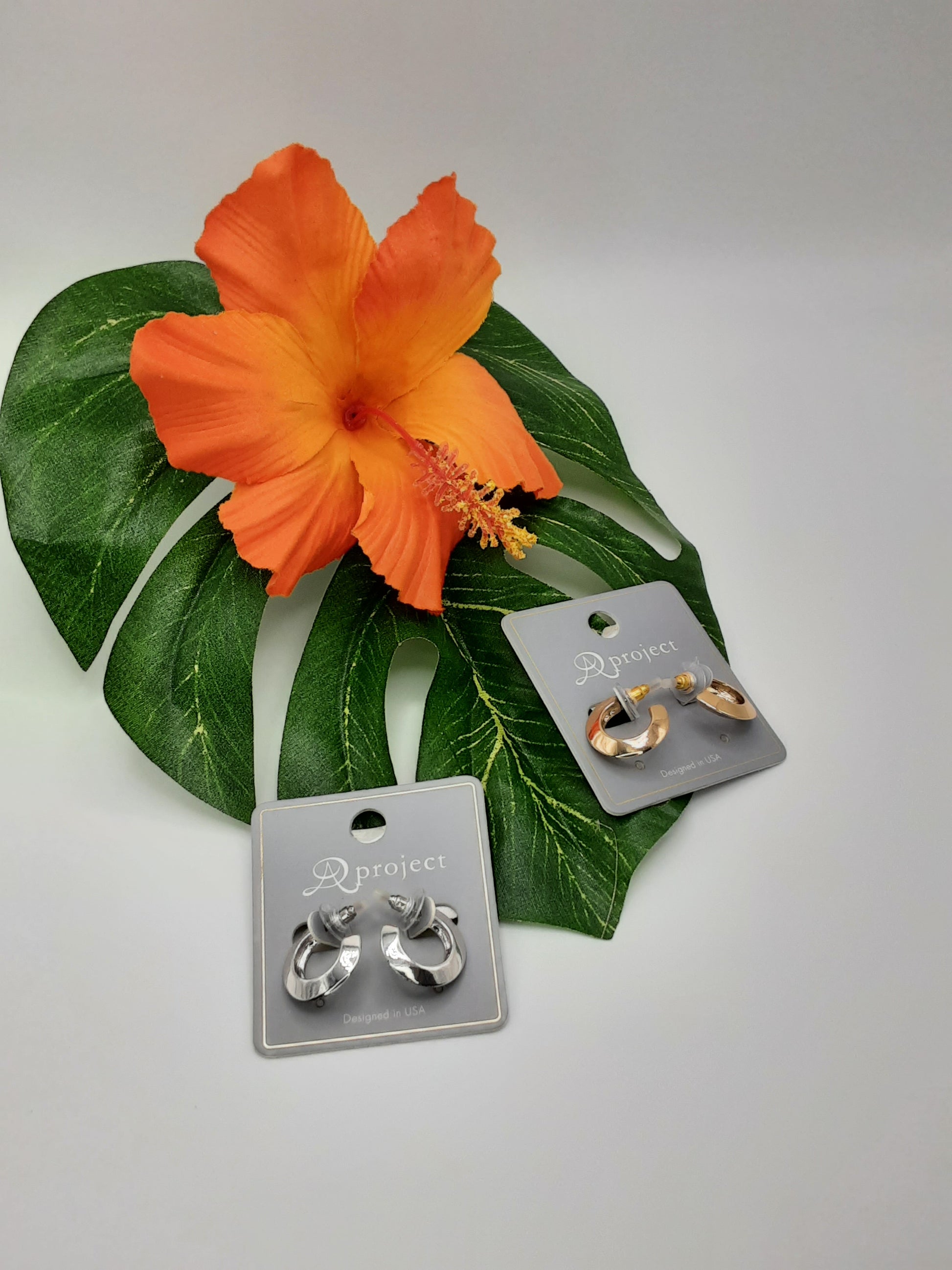 APROJECT 3796 HOOP earrings by SPECIAL EFFECTS, available in gold and silver, are showcased on cards featuring a large orange hibiscus flower and green leaf in the background.