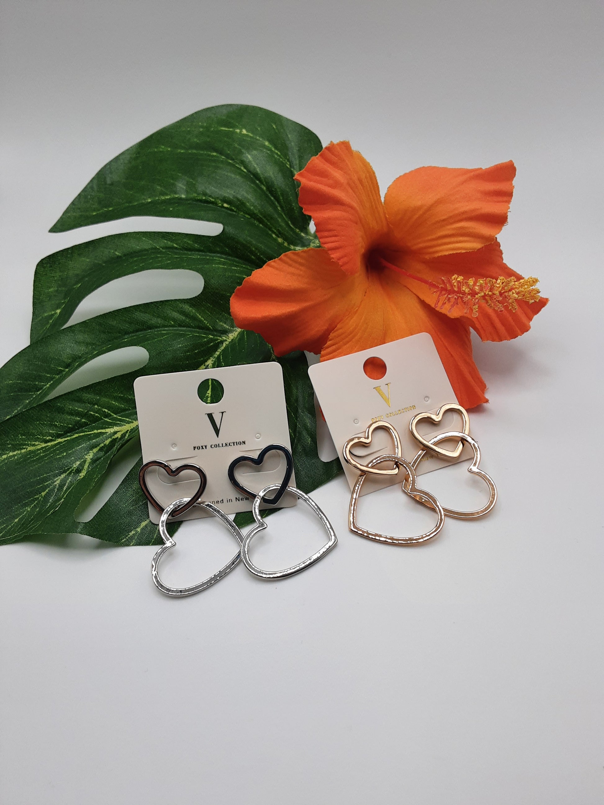 Here is a description of the product:

"Two pairs of FOXY VER4750 HEART earrings from the SPECIAL EFFECTS brand, available in silver and gold, displayed on cards with a large orange flower and green leaves in the background.