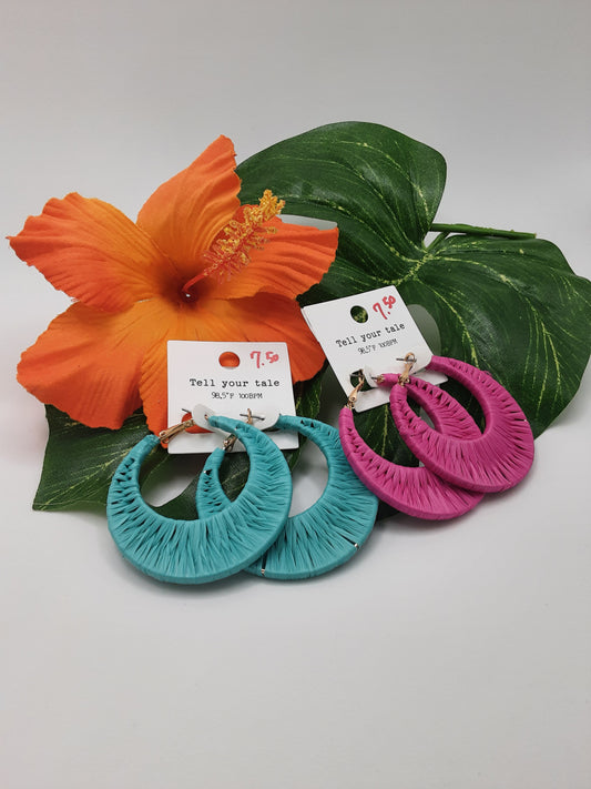 Two pairs of SPECIAL EFFECTS Raffia Wrapped Hoop Earrings, one in turquoise and one in white hot pink, are displayed in front of an orange hibiscus flower and green leaves. Each pair is priced at $9.99.