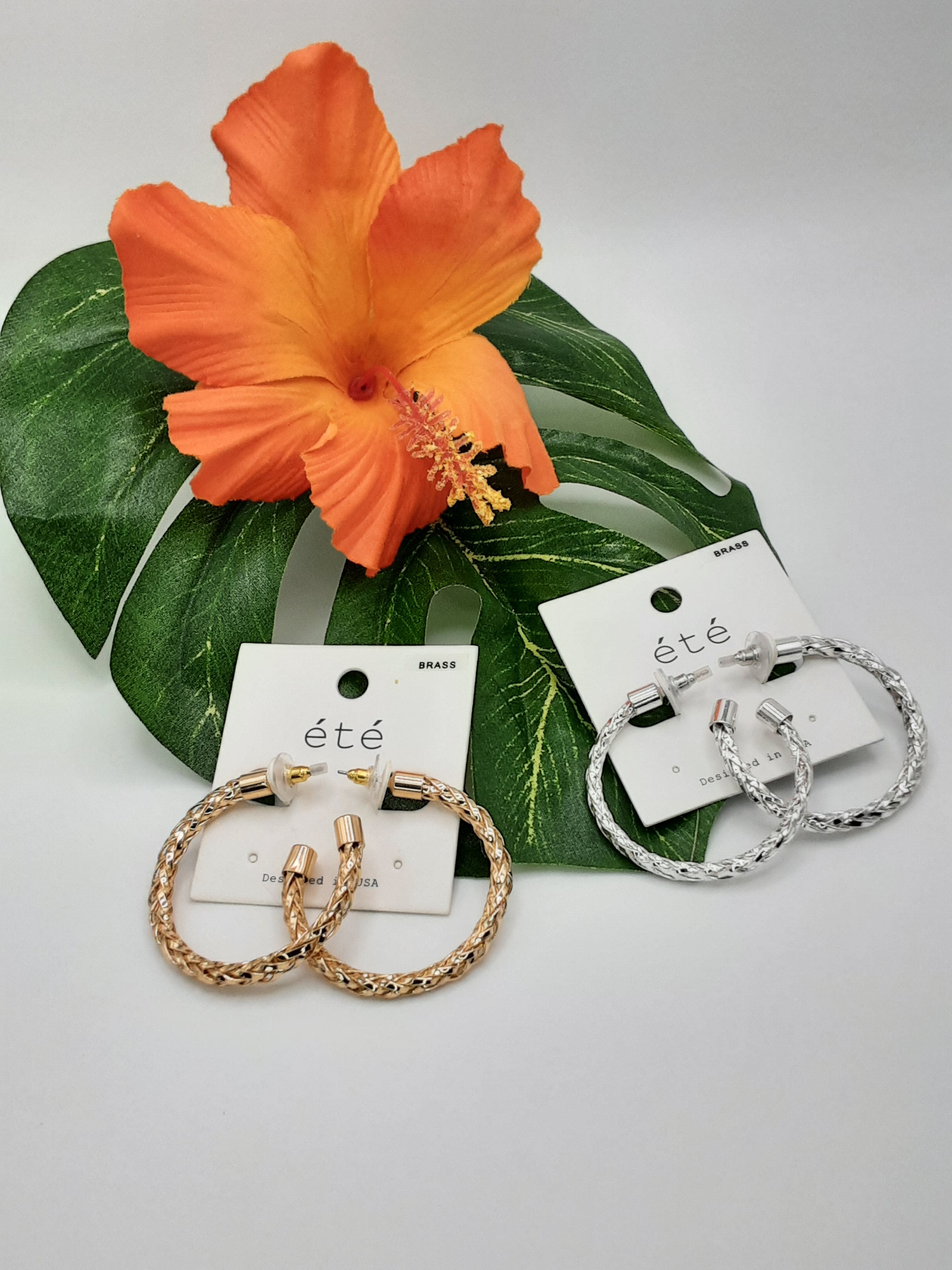 Two pairs of SPECIAL EFFECTS Woven Look Hoop Earrings, one in gold and the other in silver, are displayed on cards labeled "été" with an orange hibiscus flower and green leaf in the background. Main image