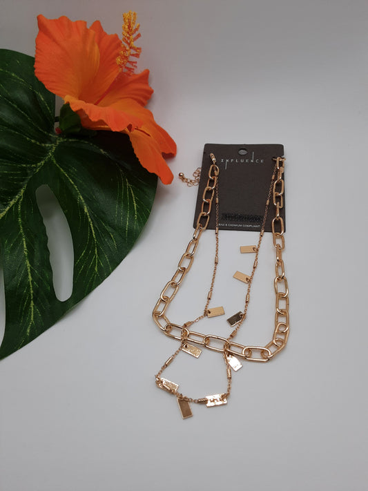 The GOLD-TONED INFLUENCE B6N7135 ASSORTED necklace and bracelet set by SPECIAL EFFECTS is elegantly presented on a display card, with a vibrant orange hibiscus flower and green leaves in the background.