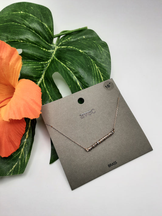 A delicate AVEC ONA481 GOLD necklace by Fashion Go, featuring a bar pendant, is displayed on a dark gray card with "avec" branding and "16 in" length noted. It lies on a green leaf with an orange flower placed beside it.