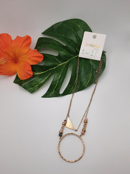 A gold necklace featuring a triangular and circular pendant, embellished with small beads, is showcased on a green leaf beside an orange flower. A tag attached to the necklace reads "Fashion Go SOUTHERN SOUL NBO193 ASSORTED.