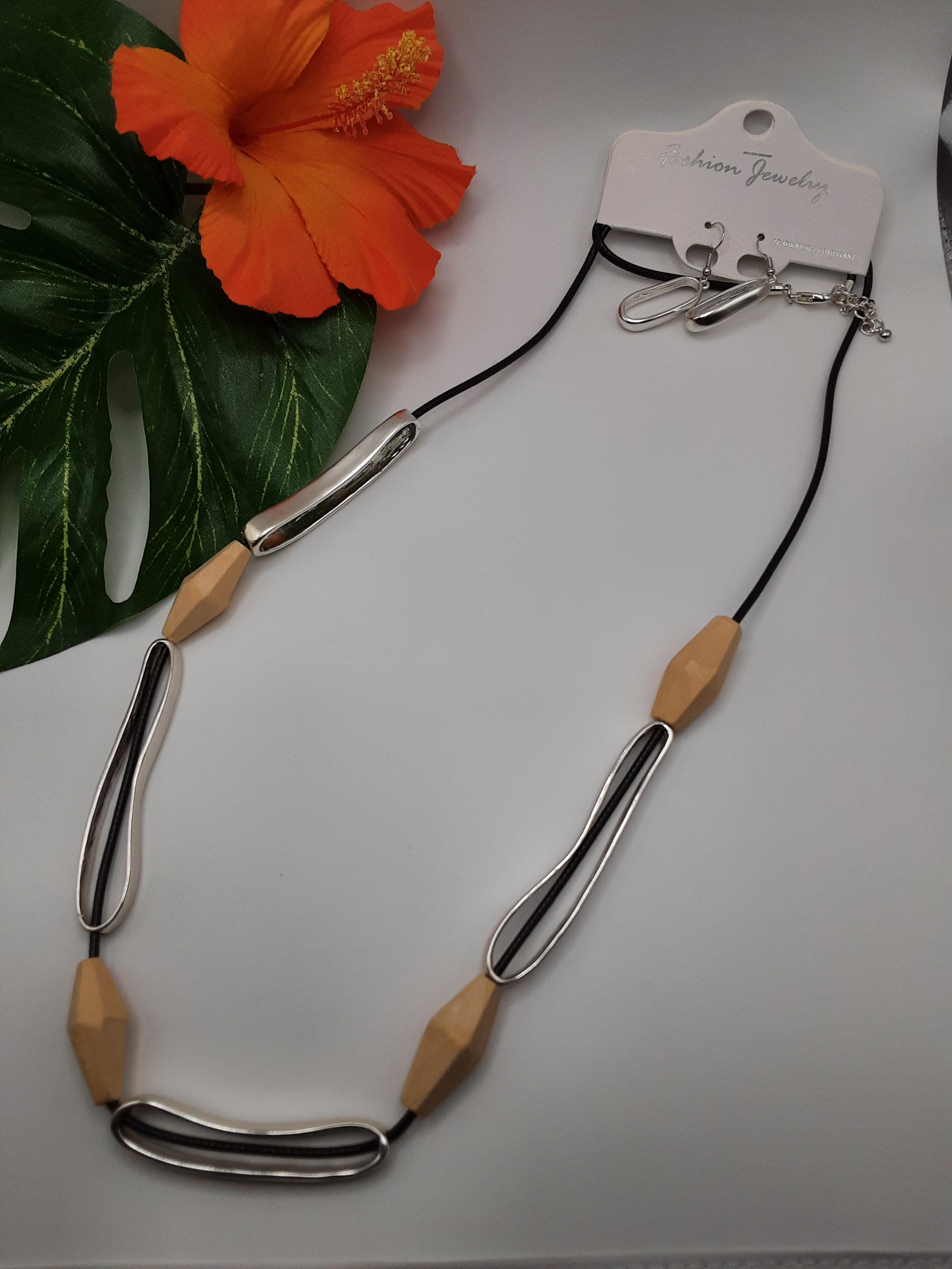 The 5 Station Natural Wood Large Bead and Black Cord Long Necklace Set by SPECIAL EFFECTS is displayed on a white surface, complemented by an orange hibiscus flower and green leaf in the background.