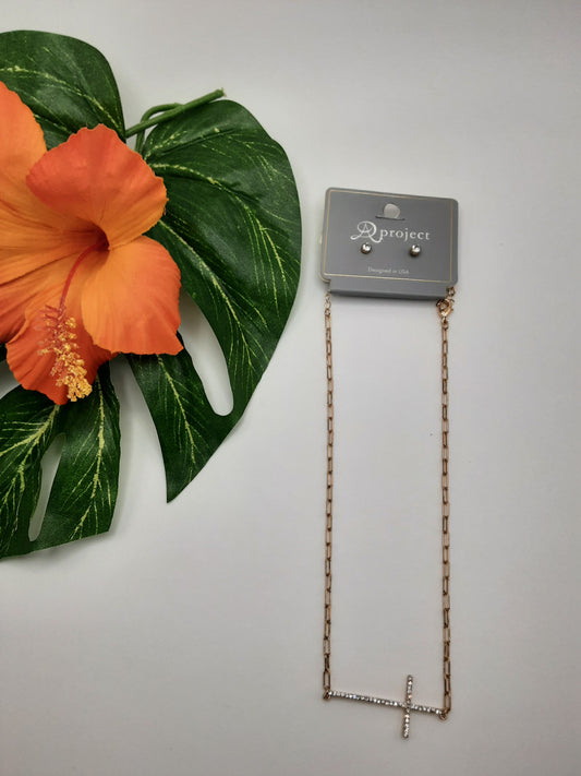 The APROJECT 3827 CROSS set, featuring a gold-tone necklace with a horizontal cross pendant and matching stud earrings from the SPECIAL EFFECTS brand, is displayed next to an orange hibiscus flower and green leaf. The set is attached to packaging labeled "A New Day.