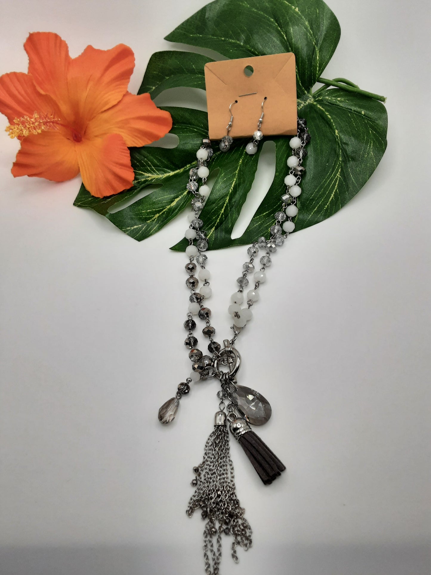 The BLANK 18116 CRYSTAL set from SPECIAL EFFECTS, featuring earrings and a necklace made of white and gray beads with a chain tassel and pendant, is elegantly displayed on green leaves next to an orange hibiscus flower.