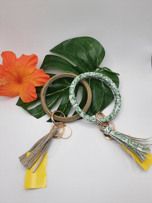 Two LAUREN LANE KEY CHAIN ASSORTED keychains from Fashion Go, one with an attached gold tassel and the other wrapped in green patterned fabric, are laid on a large green leaf. An orange hibiscus flower is placed to the left for decoration.
