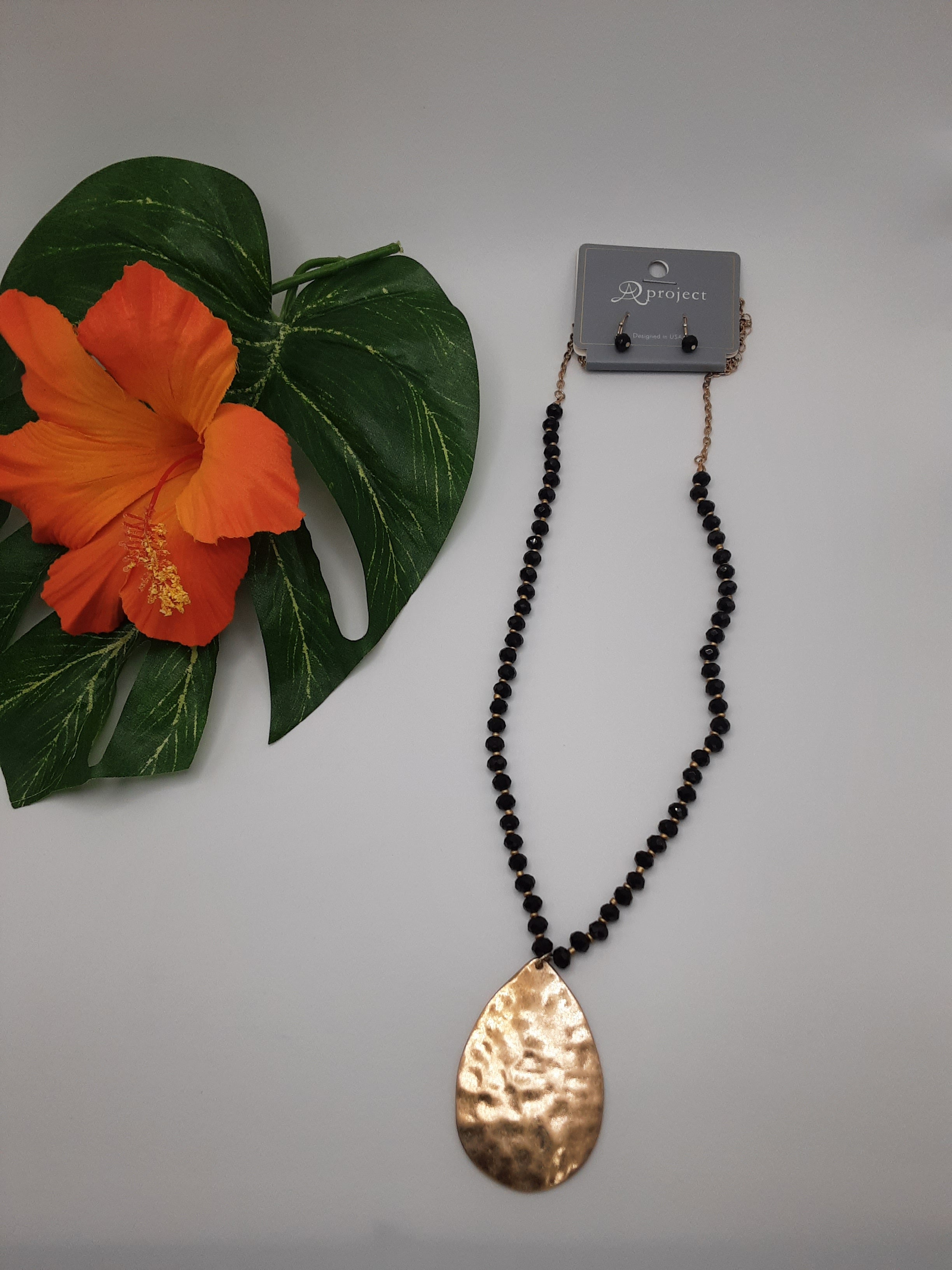 The Teardrop Pendant and Crystal Necklace Assorted from SPECIAL EFFECTS, featuring a long chain adorned with assorted bead colors and a large teardrop-shaped gold pendant, is displayed beside an orange hibiscus flower and a green leaf. Main image