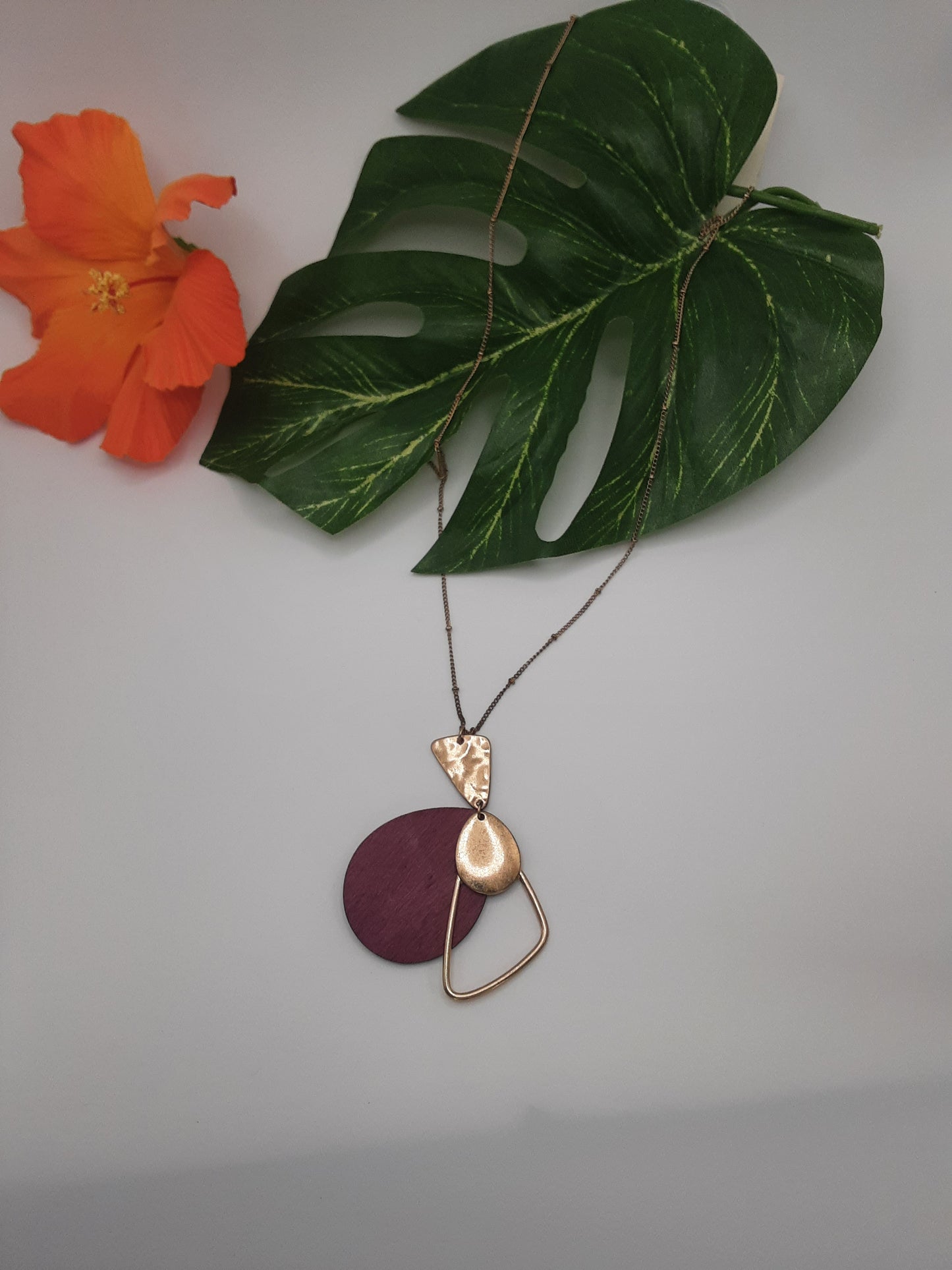 The NECK/9018 necklace by Ganz Inc, featuring two pendants—one gold and the other purple—is displayed on a white surface alongside a green leaf and an orange flower.