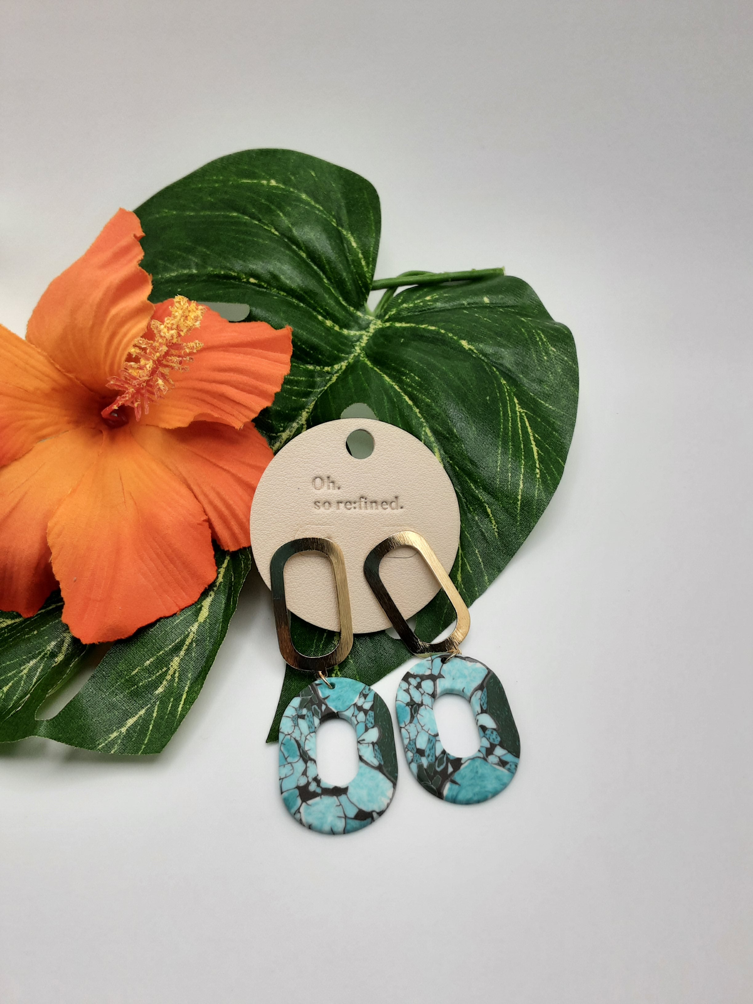 Two pairs of earrings on a tag labeled "Oh, so refined." The earrings are displayed against large green leaves and an orange hibiscus flower. One pair, from the product line OH SO ED1628MU TURQUOISE by Fashion Go, has turquoise and black patterns, while the other pair is gold-toned. Main image