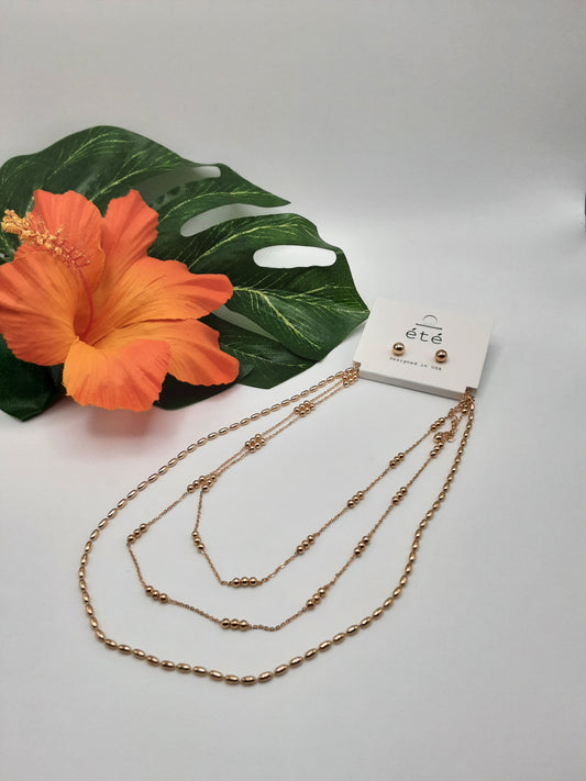 A set, displayed on a white background with a large orange hibiscus flower and green leaves as accents, includes the SPECIAL EFFECTS 3 Row Gold Bead Layered Chain Necklace and matching stud earrings, featuring a dainty gold chain necklace to complete the look.