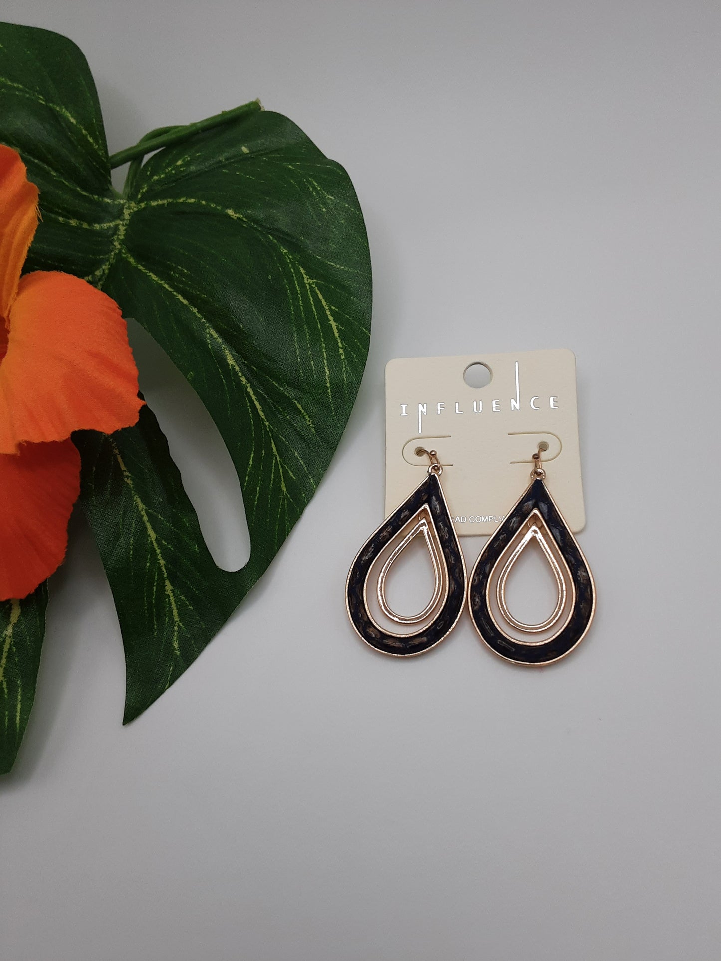 A pair of INFLUENCE B6E2615 TEARDROP earrings with a black and gold design, displayed on a SPECIAL EFFECTS branded card next to an orange flower and green leaf.