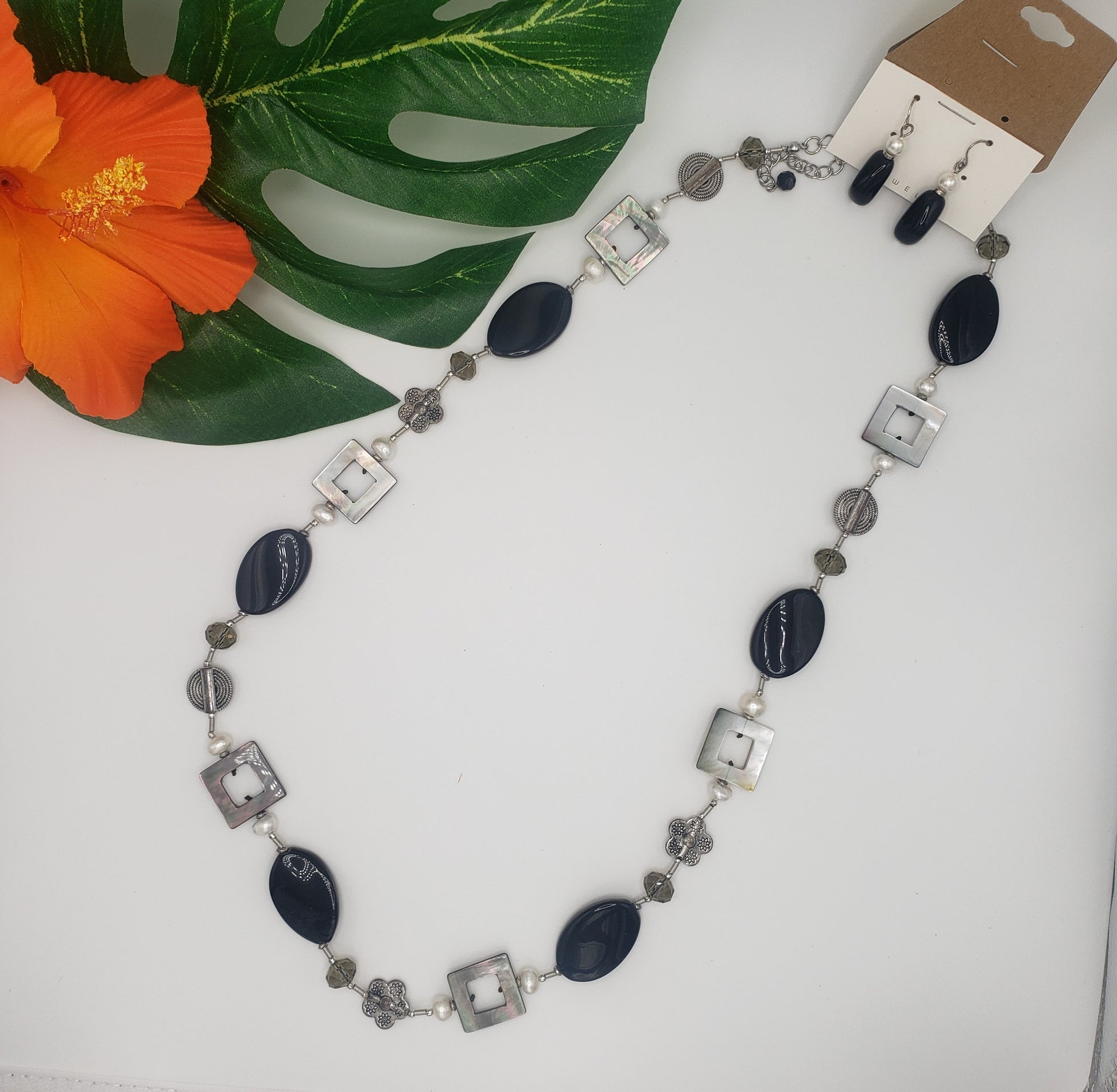 An elegant SPECIAL EFFECTS Silver-Toned Shell and Black Oval Station necklace with alternating black and white pendants, complemented by intricate silver details and matching earrings, displayed with a tropical leaf and flower.