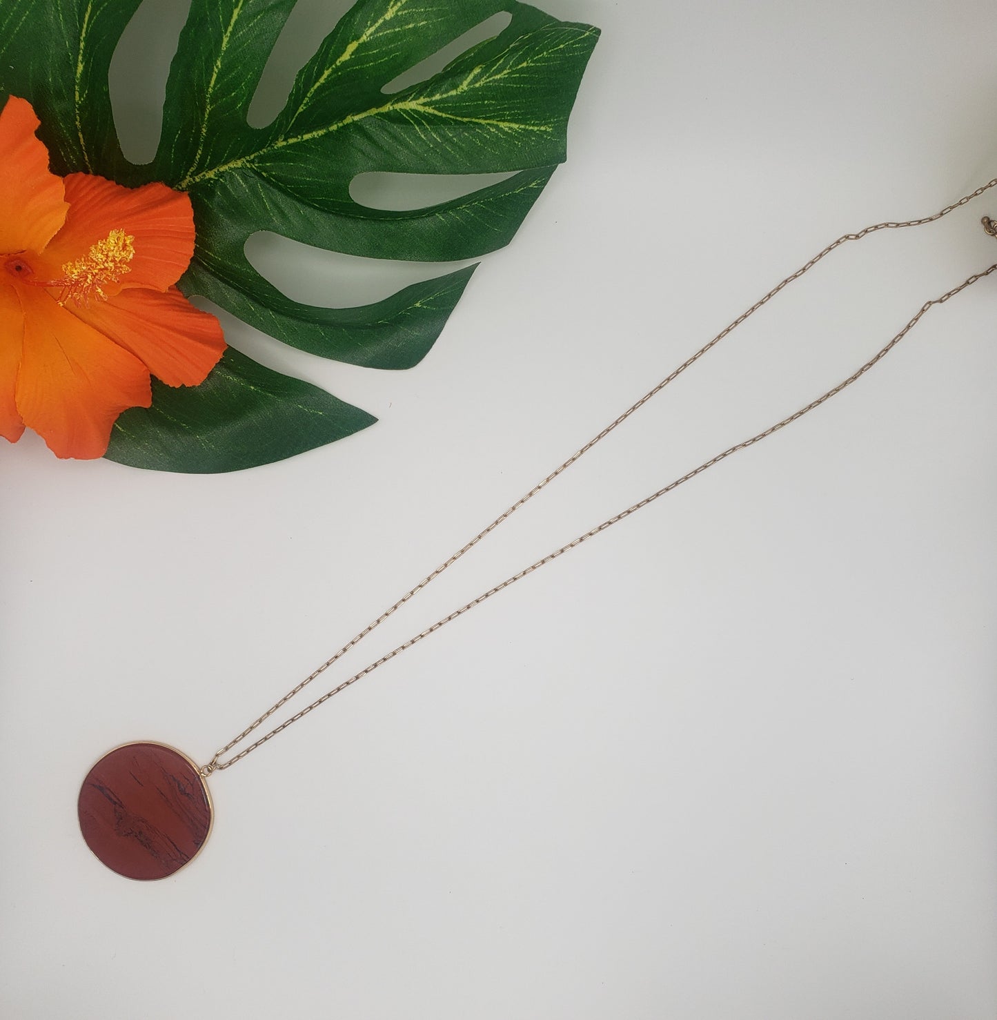 The Natural Stone Disc Necklace in rust by Garden Party is beautifully displayed against a white background, accompanied by a tropical leaf and an orange flower.